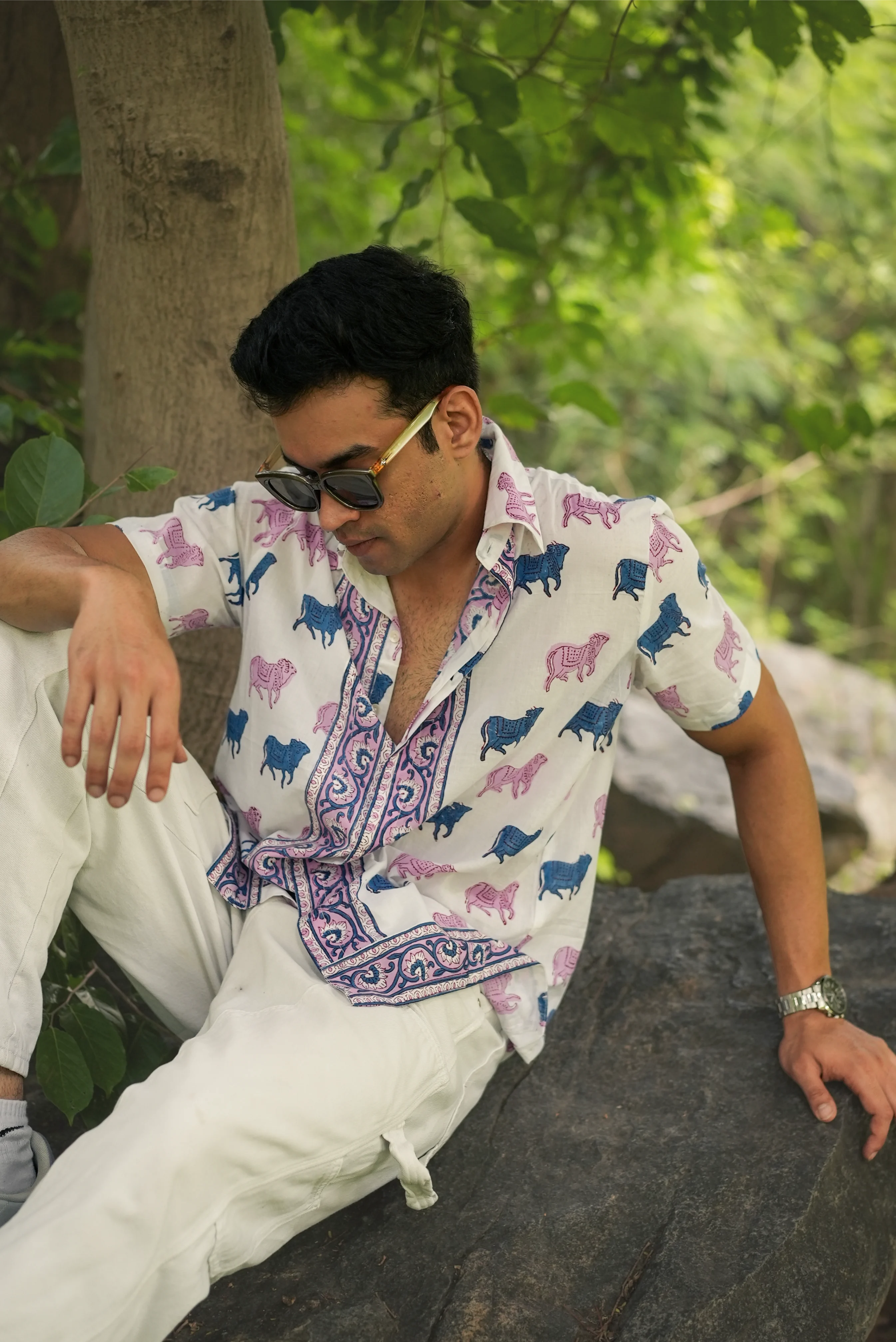 Man wearing a colorful Sattva and Sage shirt with animal patterns and white pants, sitting outdoors.