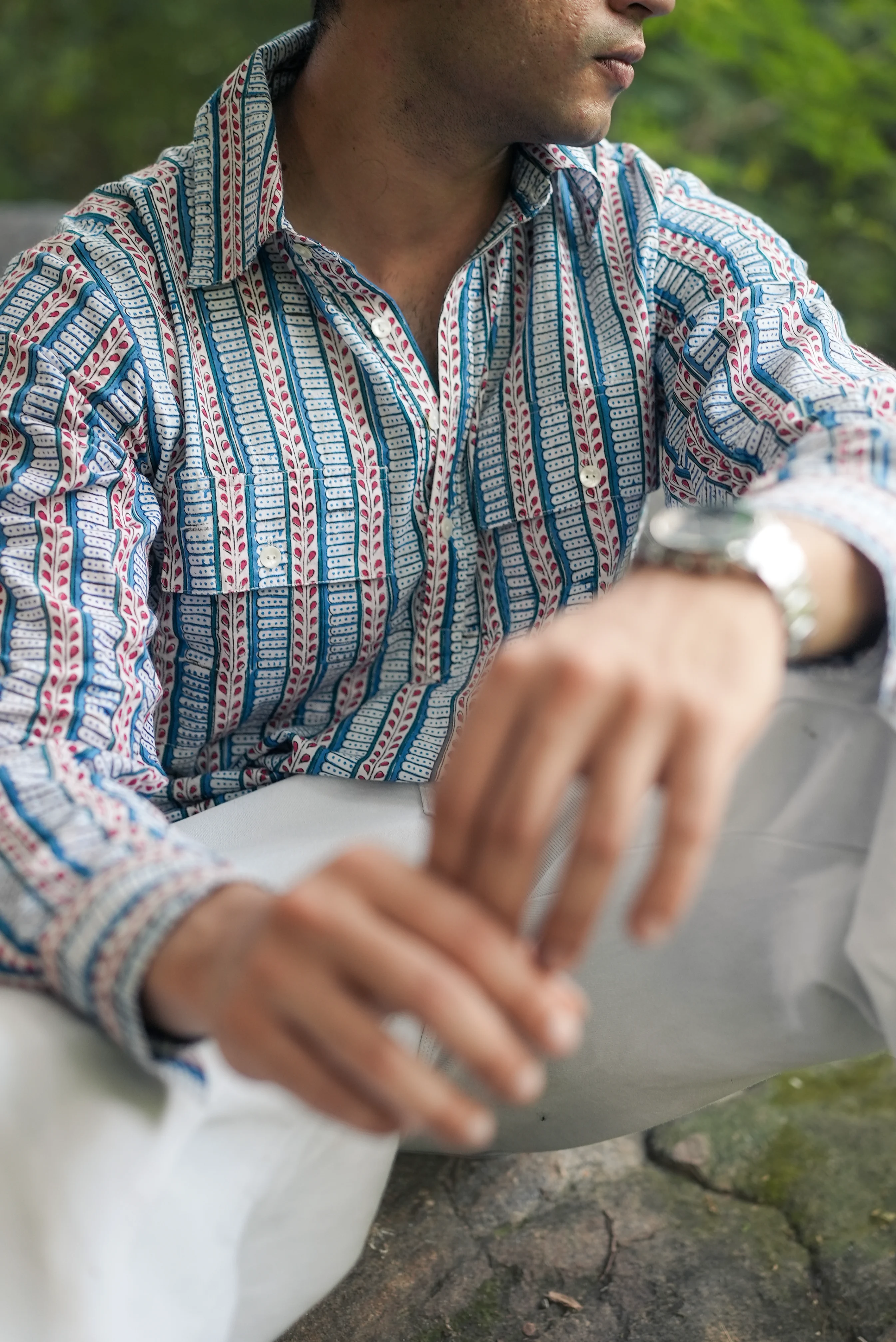 Person wearing a patterned Sattva and Sage shirt sitting outdoors on a stone surface.
