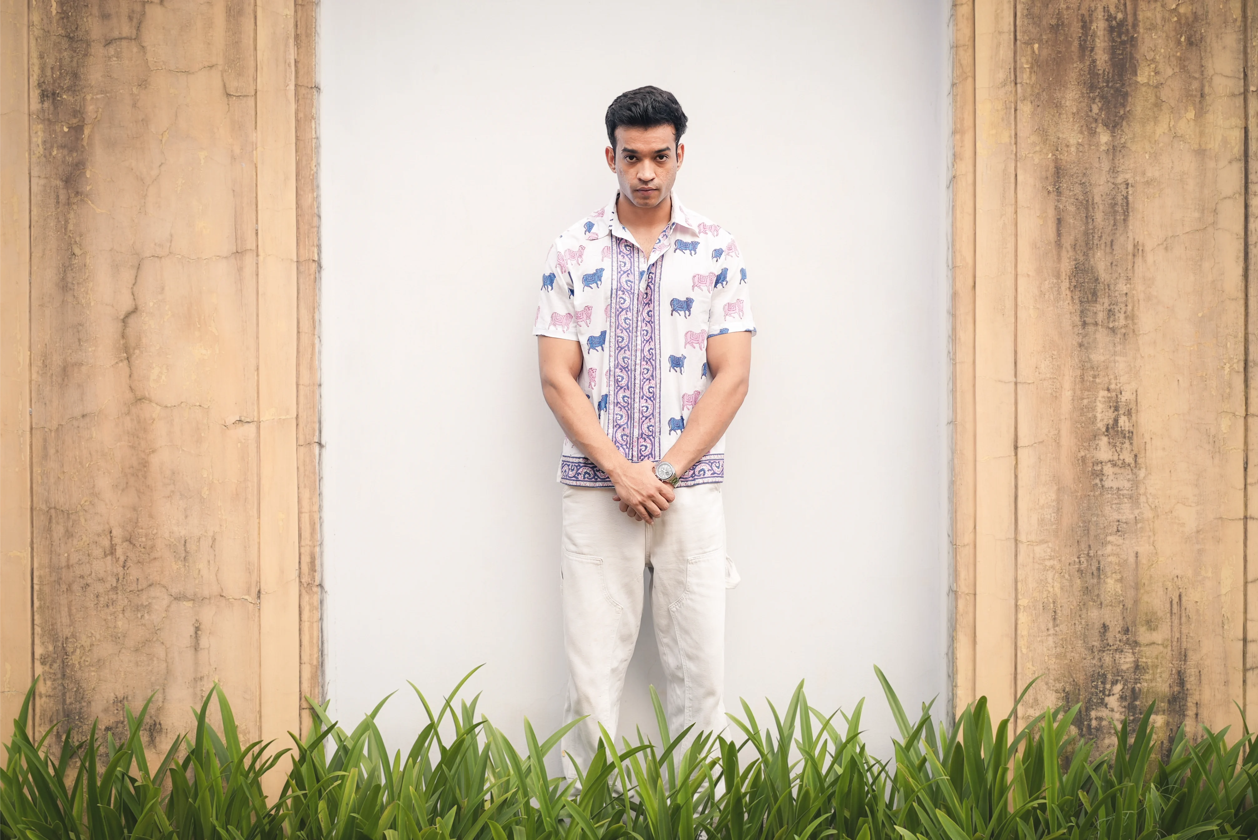 Man wearing a patterned Sattva and Sage shirt with light pants, standing in front of a white wall with wooden frames.
