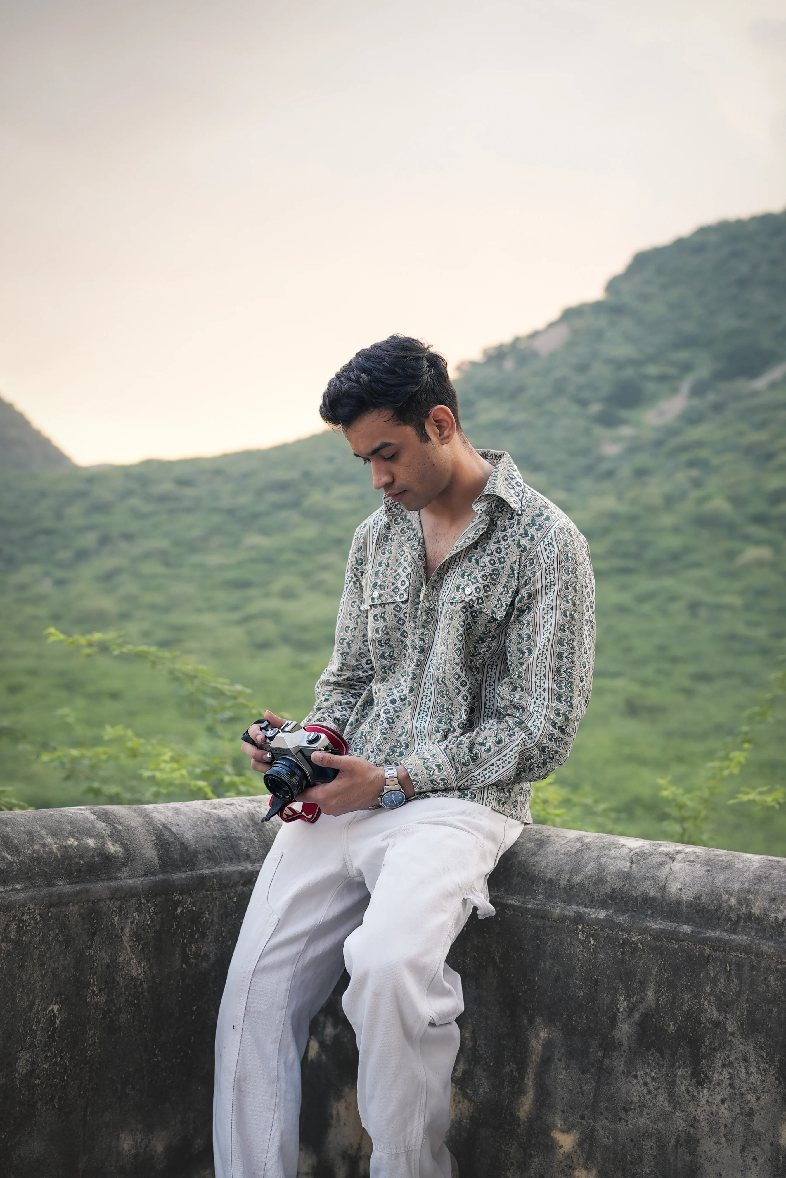 Man wearing Sattva and Sage clothing, sitting on a wall with mountains in the background.