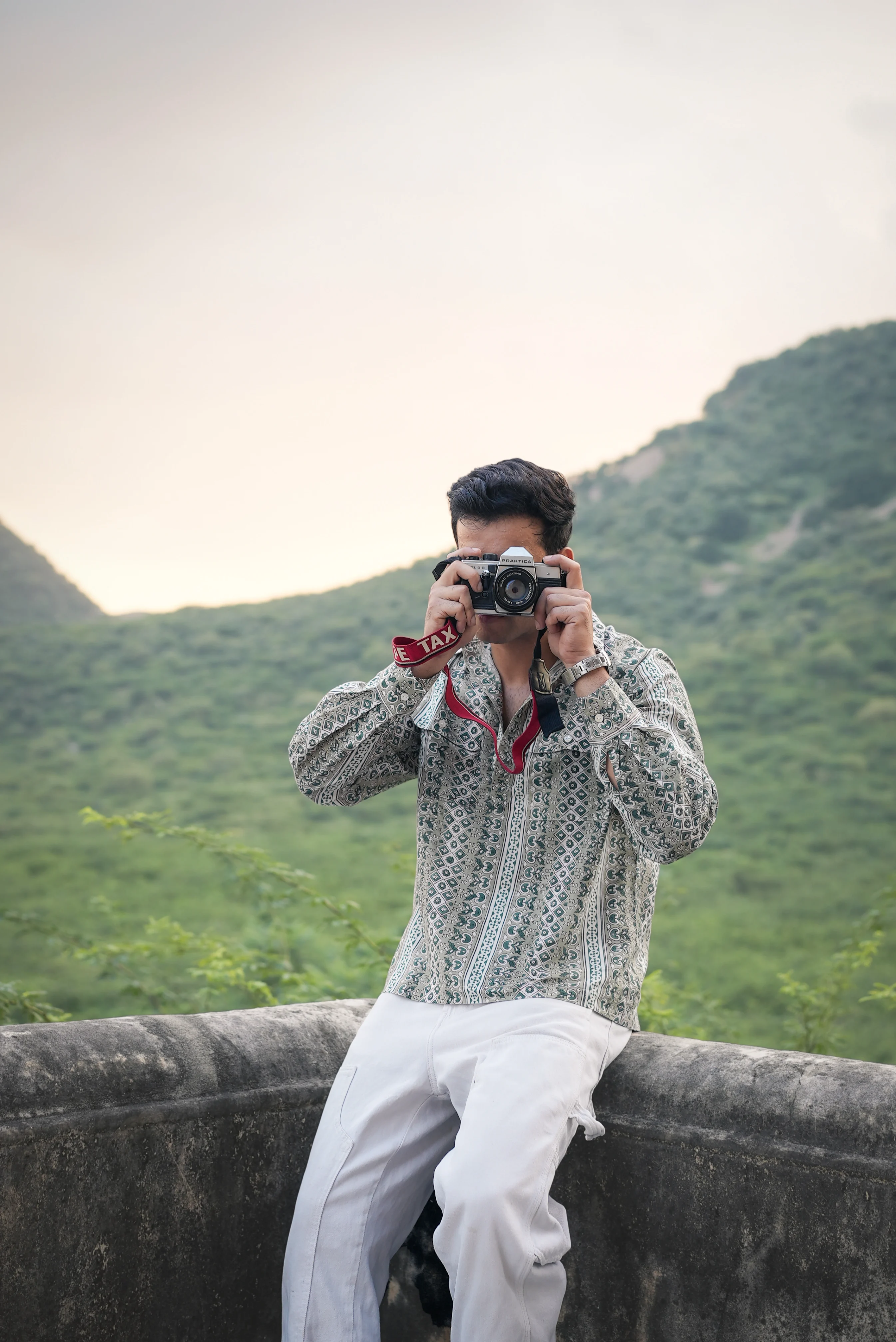 Person wearing Sattva and Sage clothing, taking a photo with a camera in a scenic outdoor setting.
